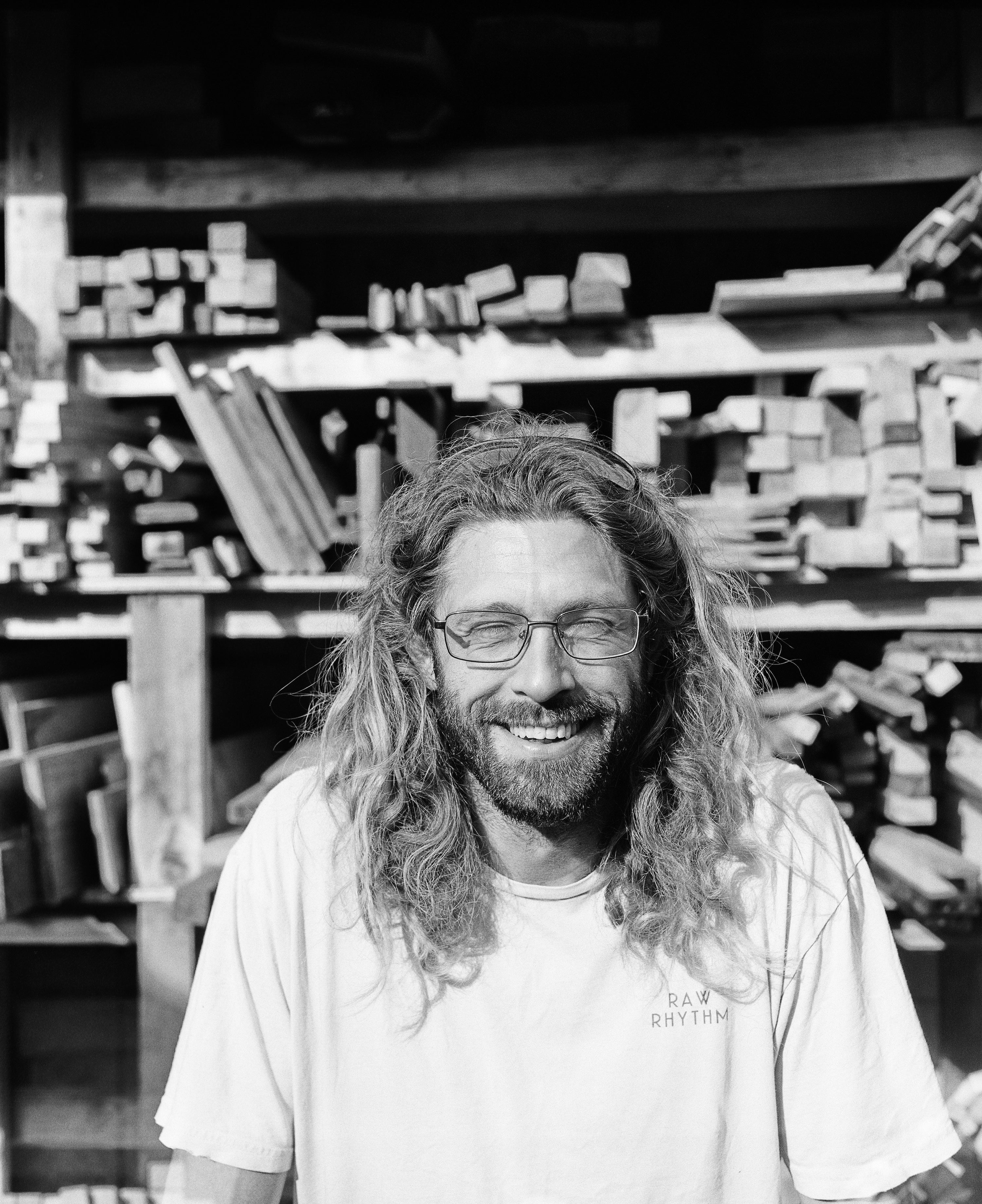 Man with long hair and beard wearing glasses and a white t-shirt in a workshop.